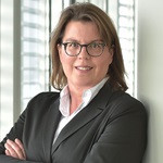 A person with long hair in a black suit standing in a modern office, with arms crossed. Portrait of Regina Wolter.