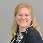 A woman with long blonde hair wearing a black and white patterned blouse, set against a grey background. Portrait of Sarah Knauber.