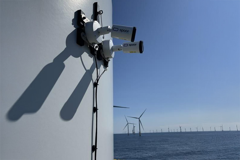 Zwei Überwachungskameras sind an einem Windkraftgenerator montiert, mit Blick auf den Ozean und Windräder im Hintergrund.