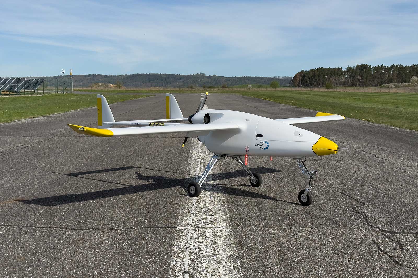 Ein weißes UAV mit gelben Akzenten steht auf einer Landebahn unter blauem Himmel und grünem Hintergrund.