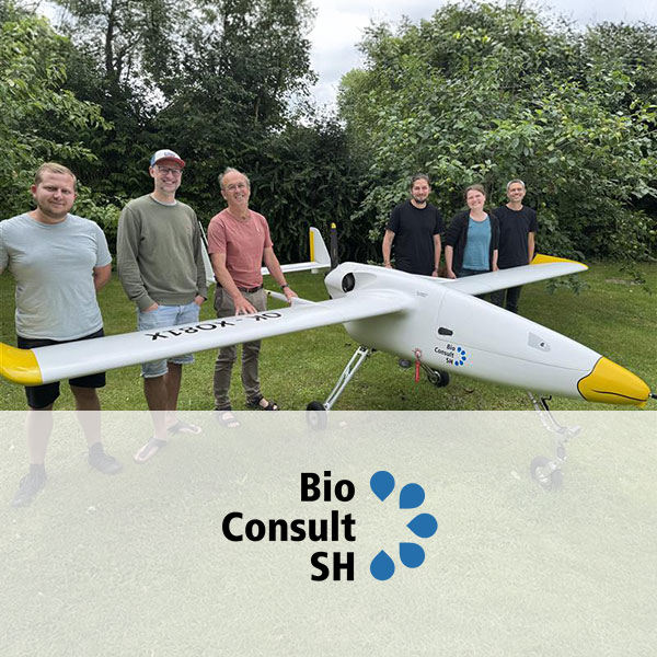 A group of people stands next to a small drone on a grassy area, surrounded by greenery and trees.