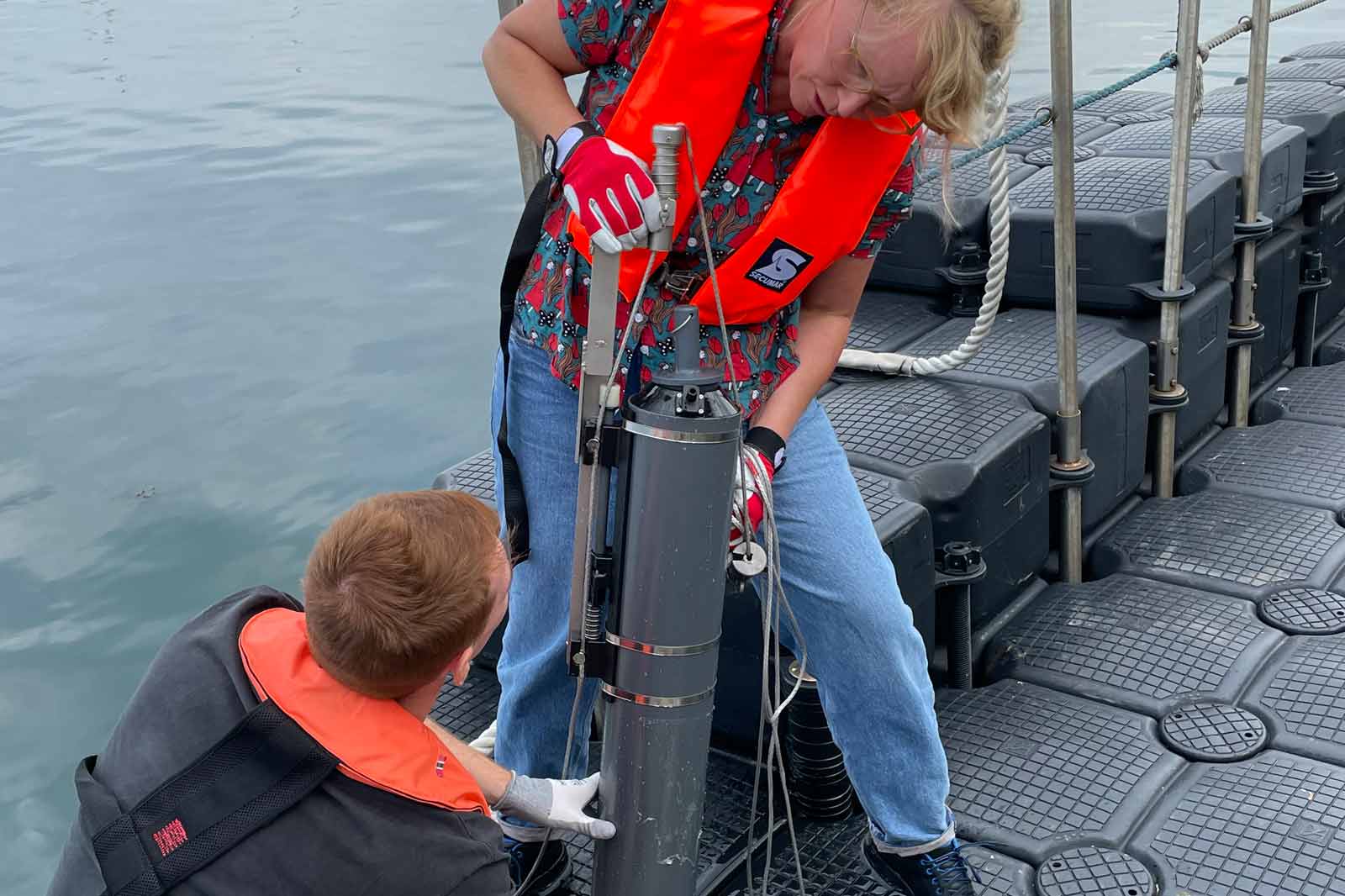 Zwei Personen auf einem Boot arbeiten zusammen an einem Wassergerät, während ein ruhiger Wasserhintergrund zu sehen ist.