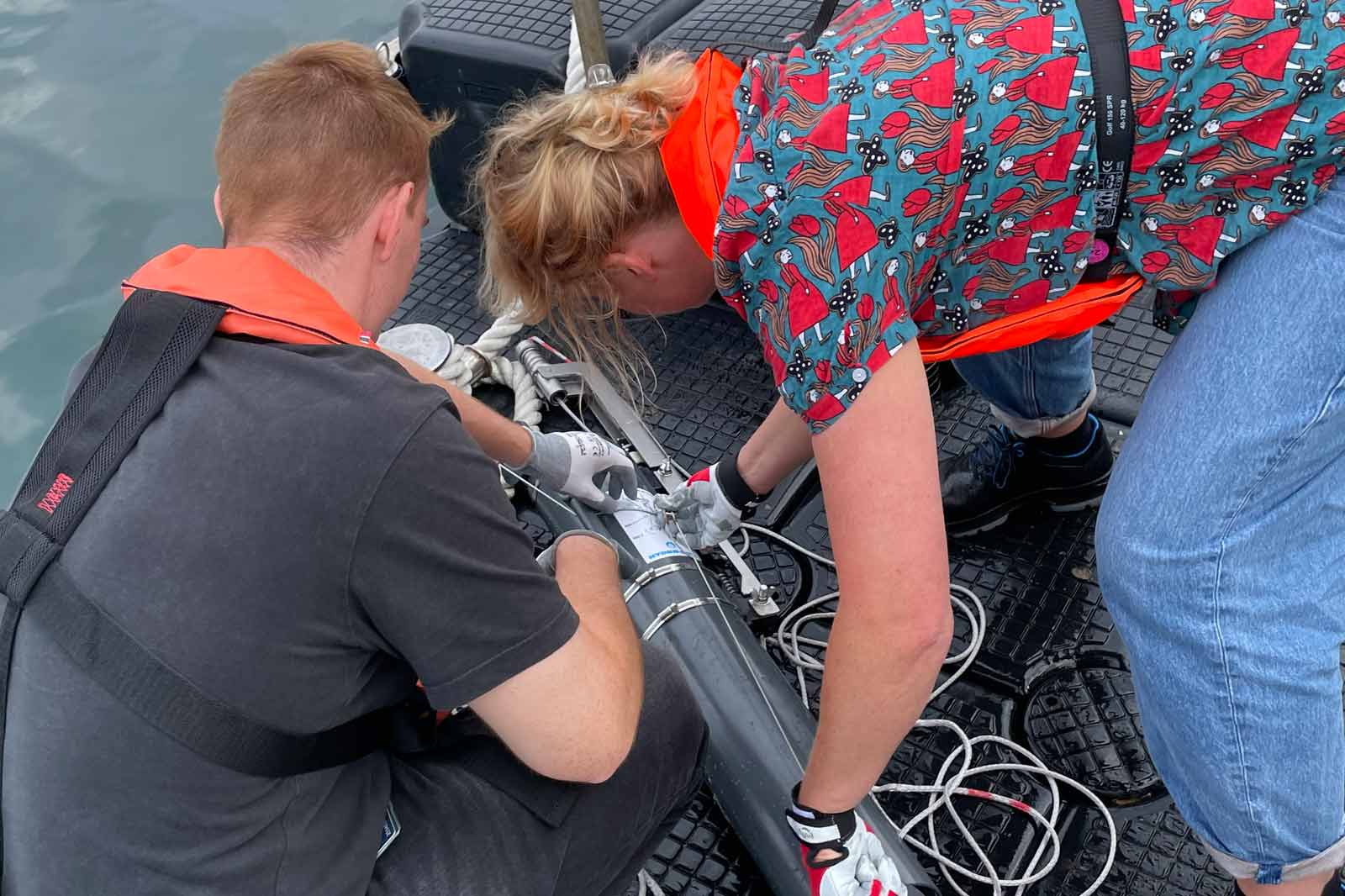 Two people are working on a device on a boat, wearing life vests and handling cables.