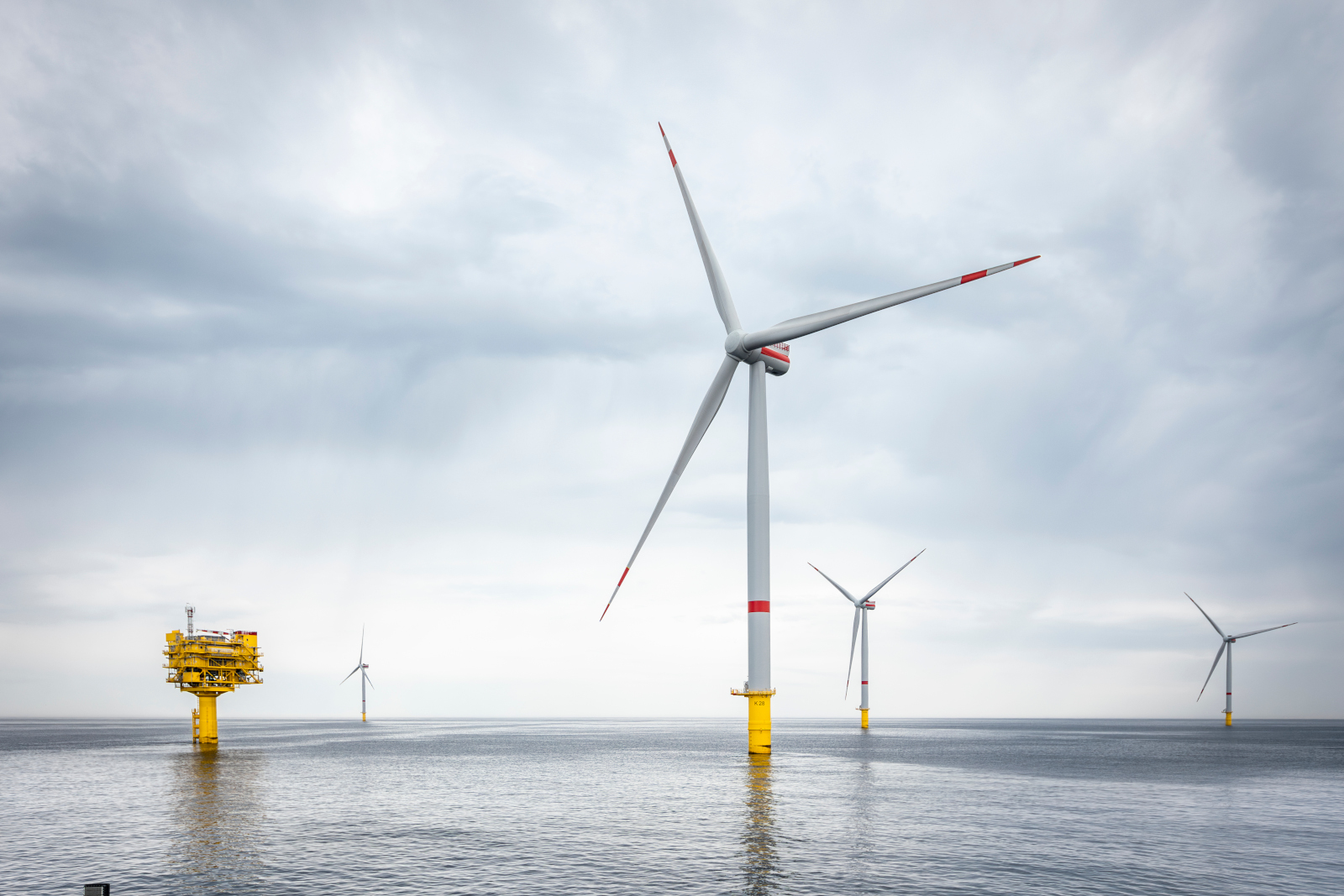 Offshore wind turbines stand in calm water under a cloudy sky. Yellow bases are visible.
