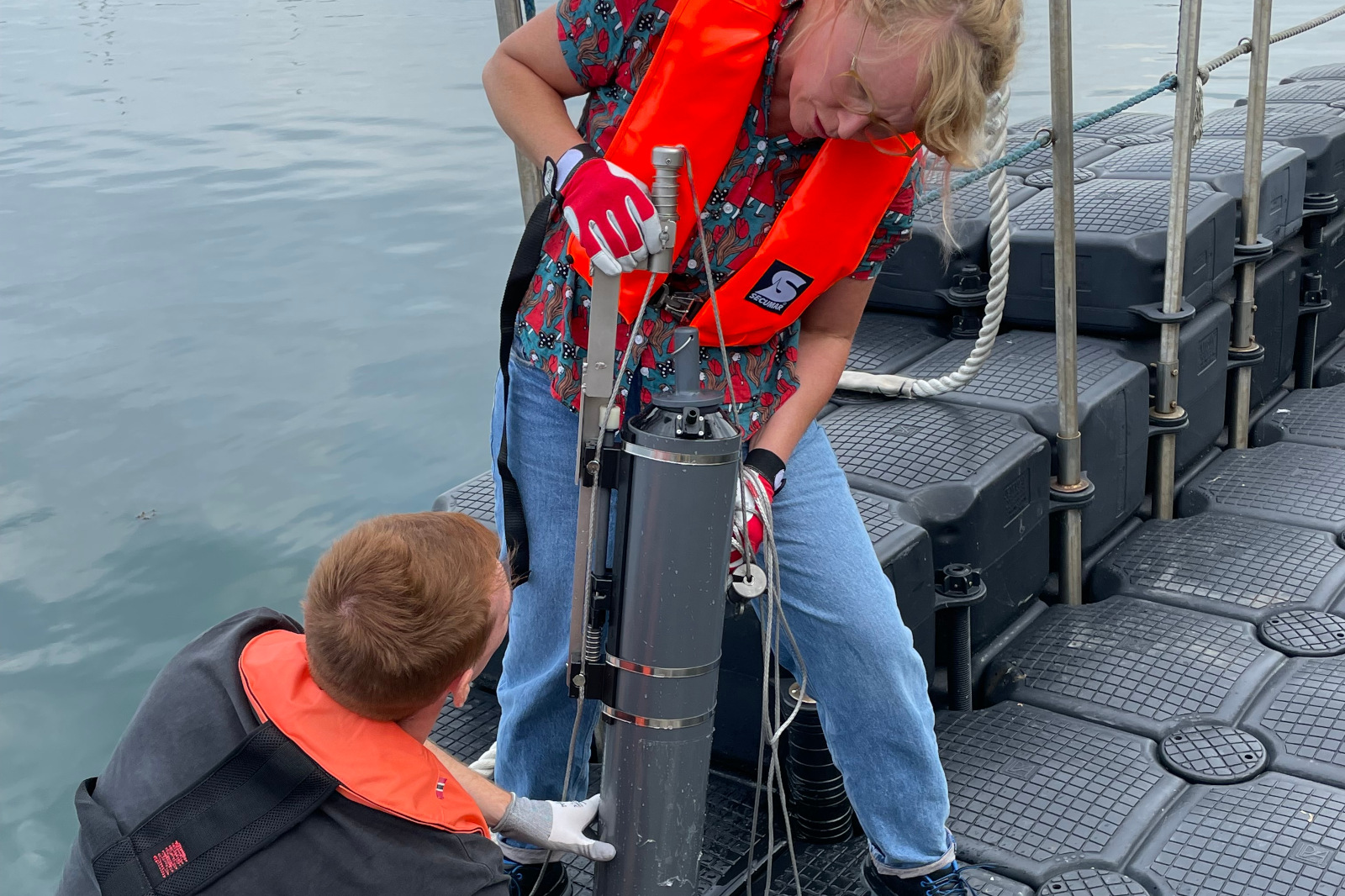 Two individuals are operating marine equipment on a dock, wearing life jackets and gloves.
