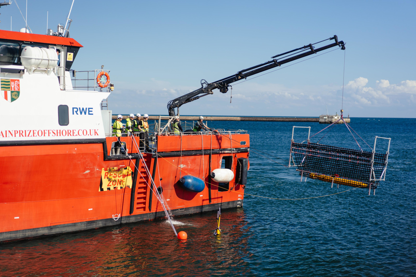 An orange research vessel lifts a net over water while workers in safety gear observe.