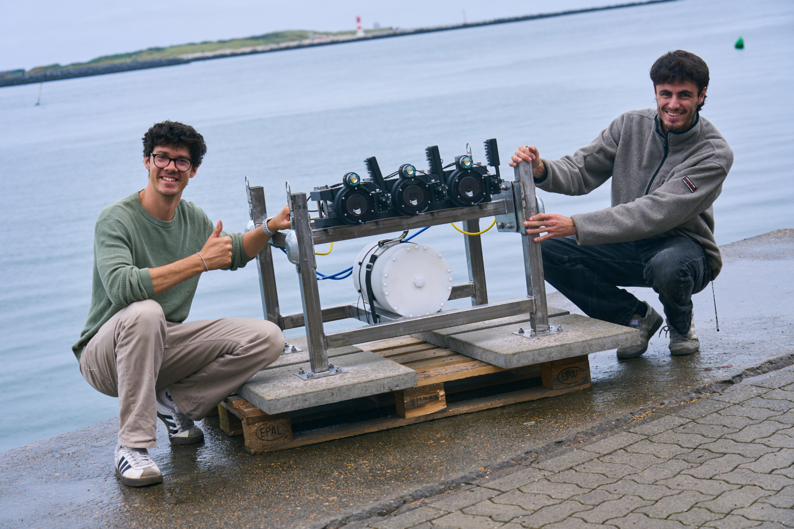 Two men showcase a technical device beside the water. The device features multiple cameras and a wheel system.
