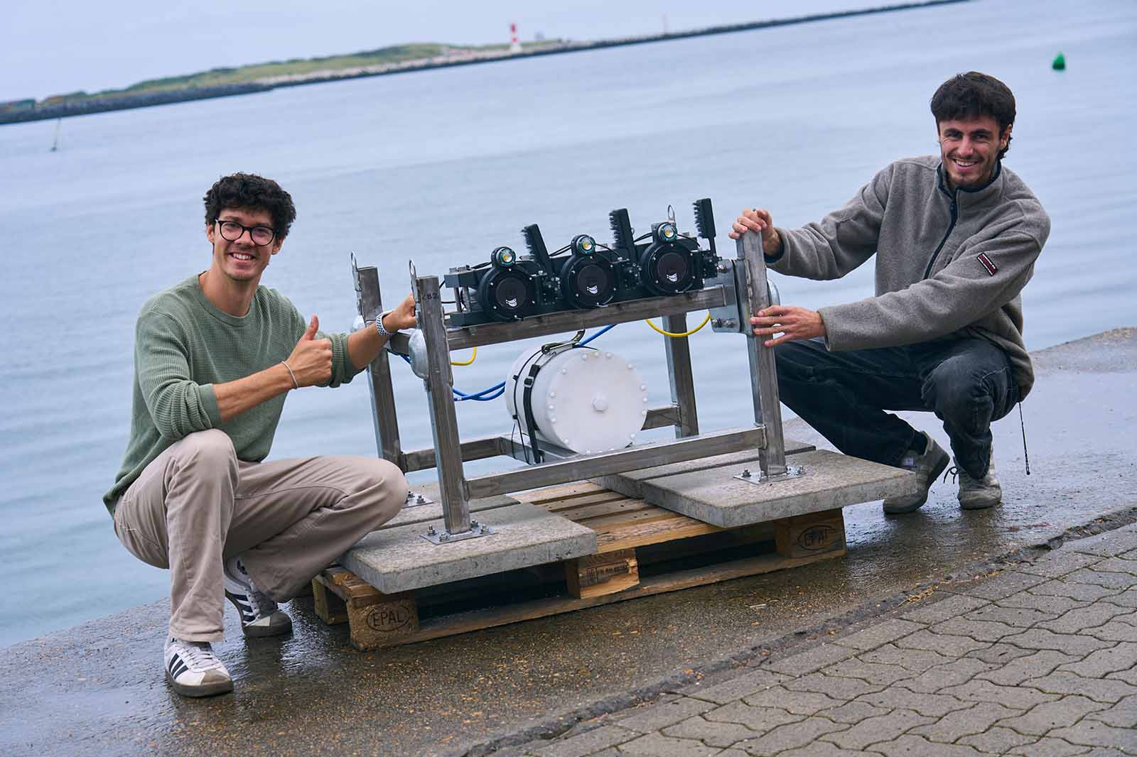 Two men kneel beside a metal frame with cameras on a pallet by the water, one giving a thumbs up.