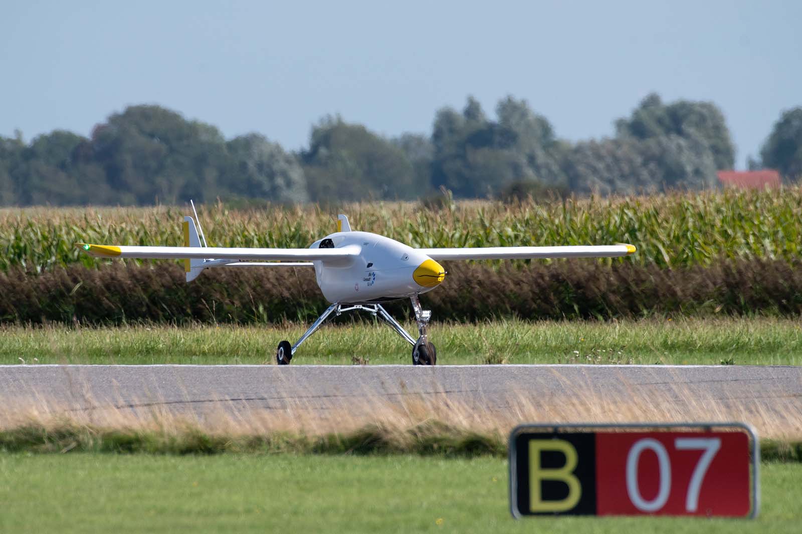 White drone with yellow nose stands on runway in front of cornfield and B 07 sign.