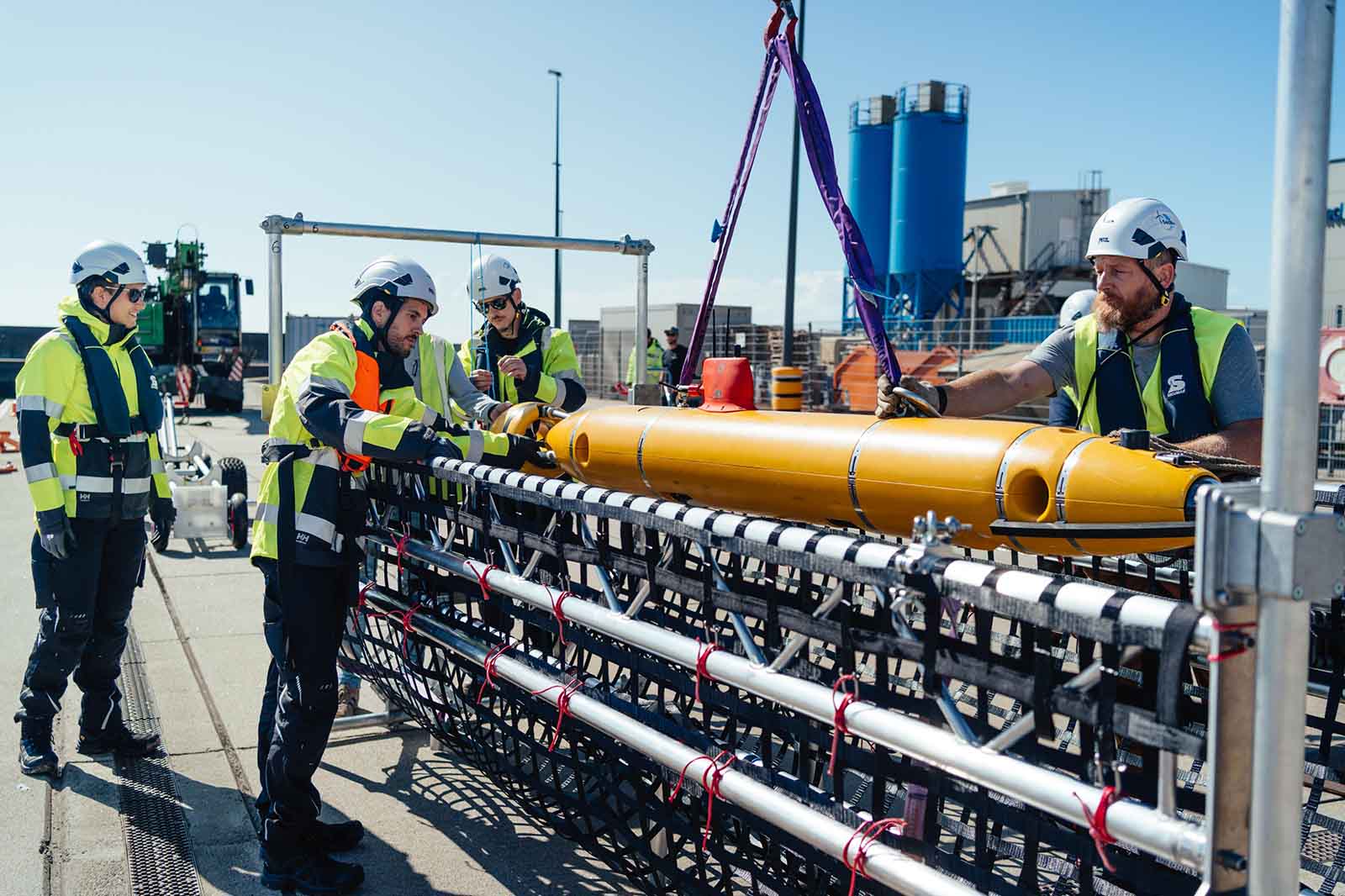 Vier Arbeiter in Schutzkleidung installieren an Deck eines Schiffes ein gelbes Gerät mit Hebegurt.