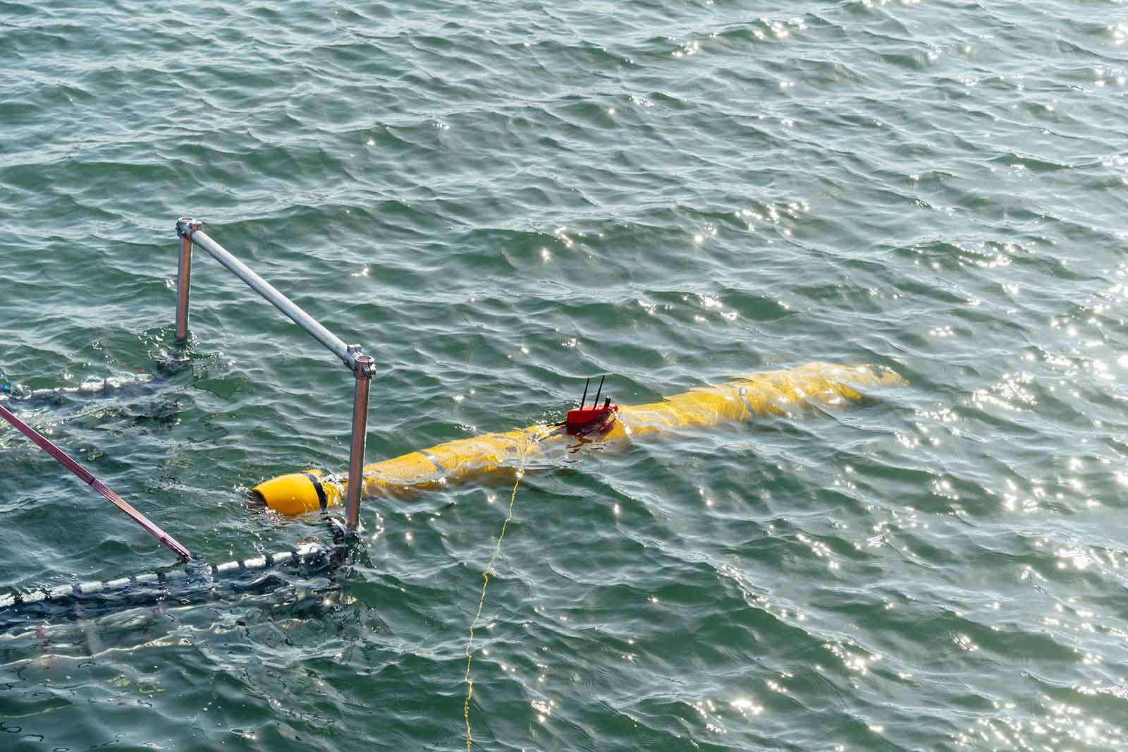 Ein gelbes Unterwasserfahrzeug mit Antennen schwimmt an der Oberfläche eines Gewässers neben einem Metallrahmen.