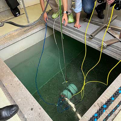Eine Person lässt ein Gerät in ein grünes Wasserbecken ab. Mehrere Kabel sind mit dem Gerät verbunden.