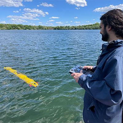 Eine Person steuert ein gelbes Boot mit einer Fernbedienung auf einem ruhigen See unter blauem Himmel.