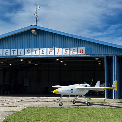 Ein kleiner gelber und weisser Flugzeugmodell steht vor einer blauen Flugzeughangar mit dem Schriftzug 'Letiště Písek'.