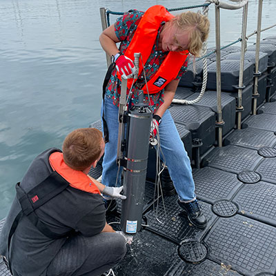 Zwei Personen in Rettungswesten arbeiten an einem Messgerät an einem Steg am Wasser.