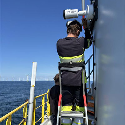 Ein Techniker steht auf einer Leiter und montiert eine Kamera an einer Offshore-Plattform vor einer Küste mit Windkraftanlagen.